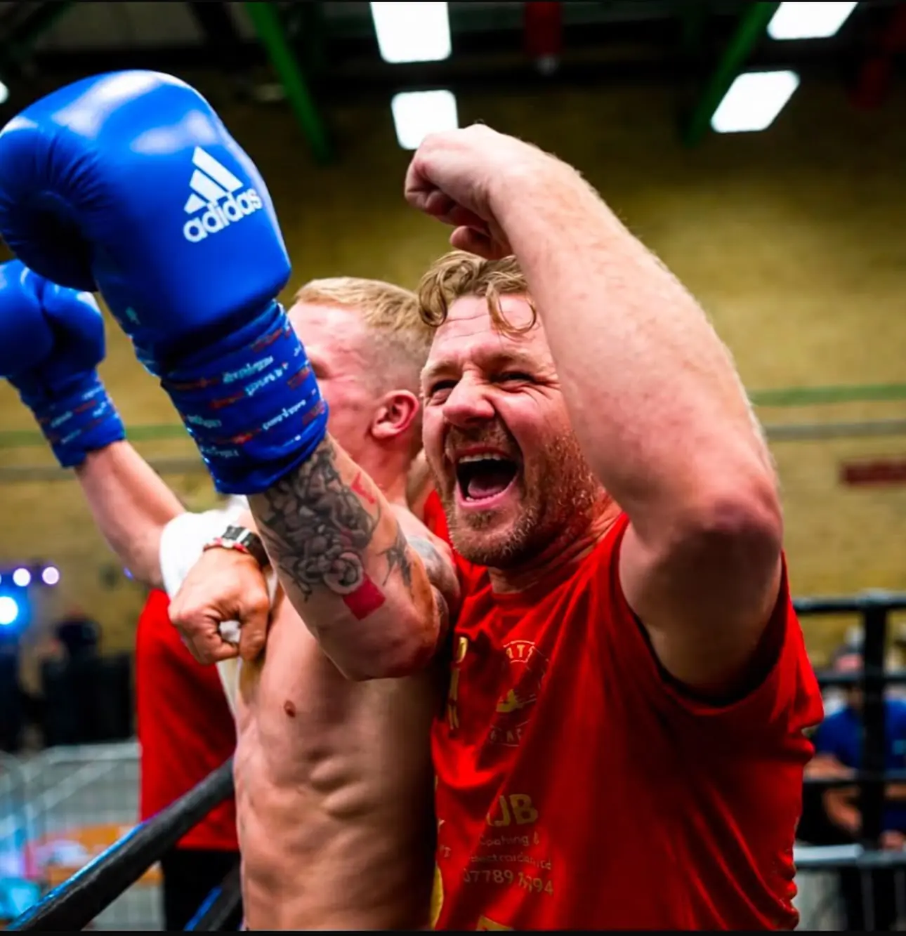A coach in a red T-shirt roaring in celebration as a boxer in blue gloves raises his arm at the ring ropes