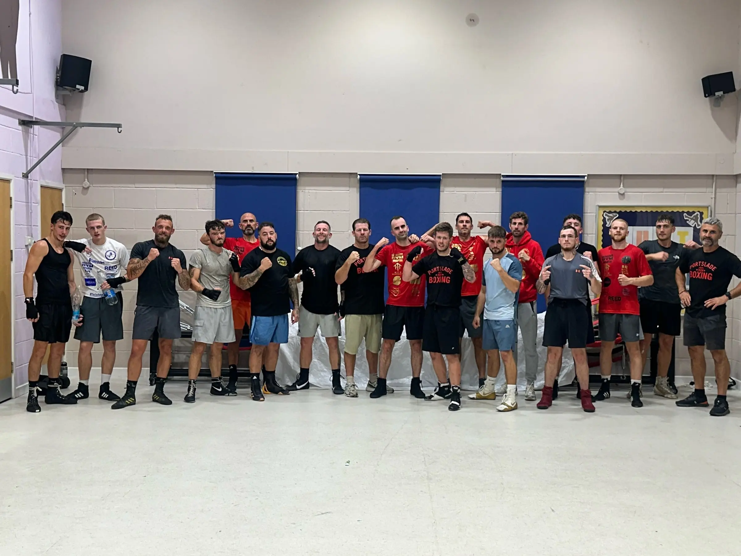 Around twenty boxers lined up in a sports hall with blue window blinds, some in red club T-shirts
