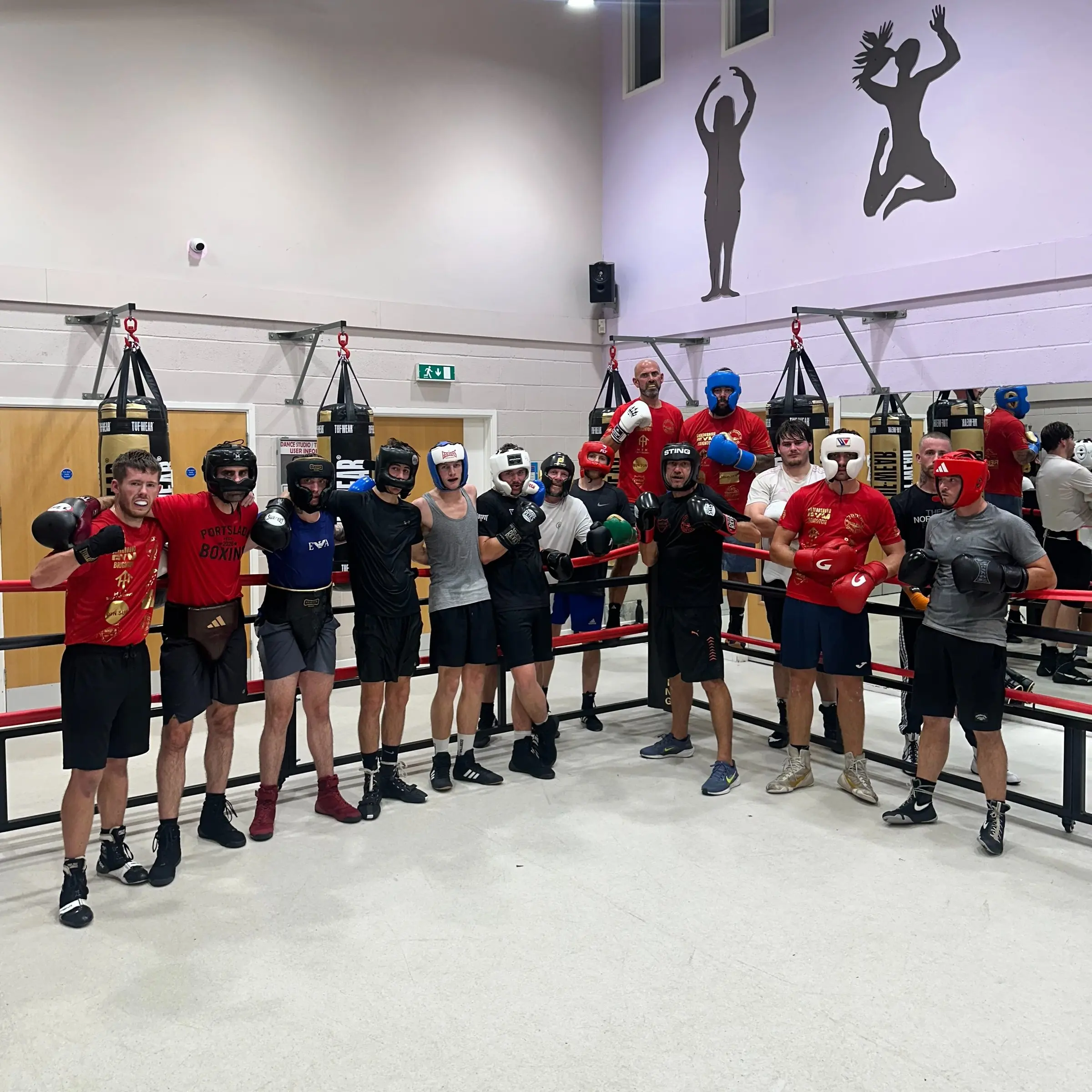 A row of boxers in head guards and gloves standing along the ropes of a ring in a bright hall