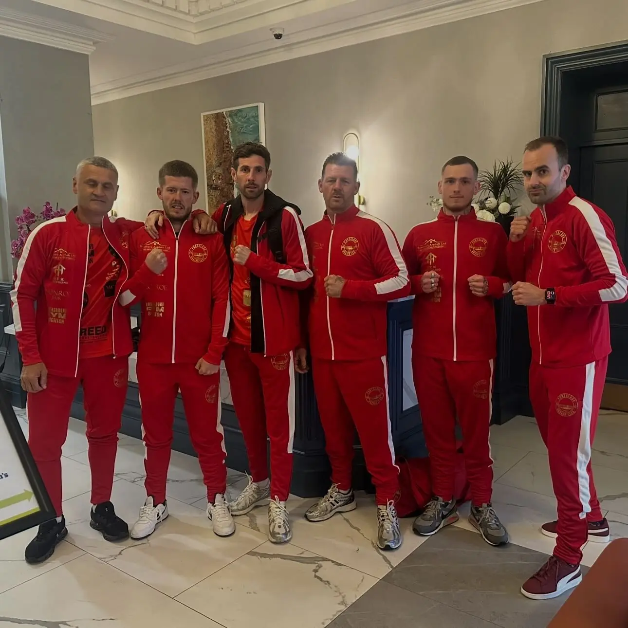 Six men in matching red club tracksuits standing together in a hotel lobby