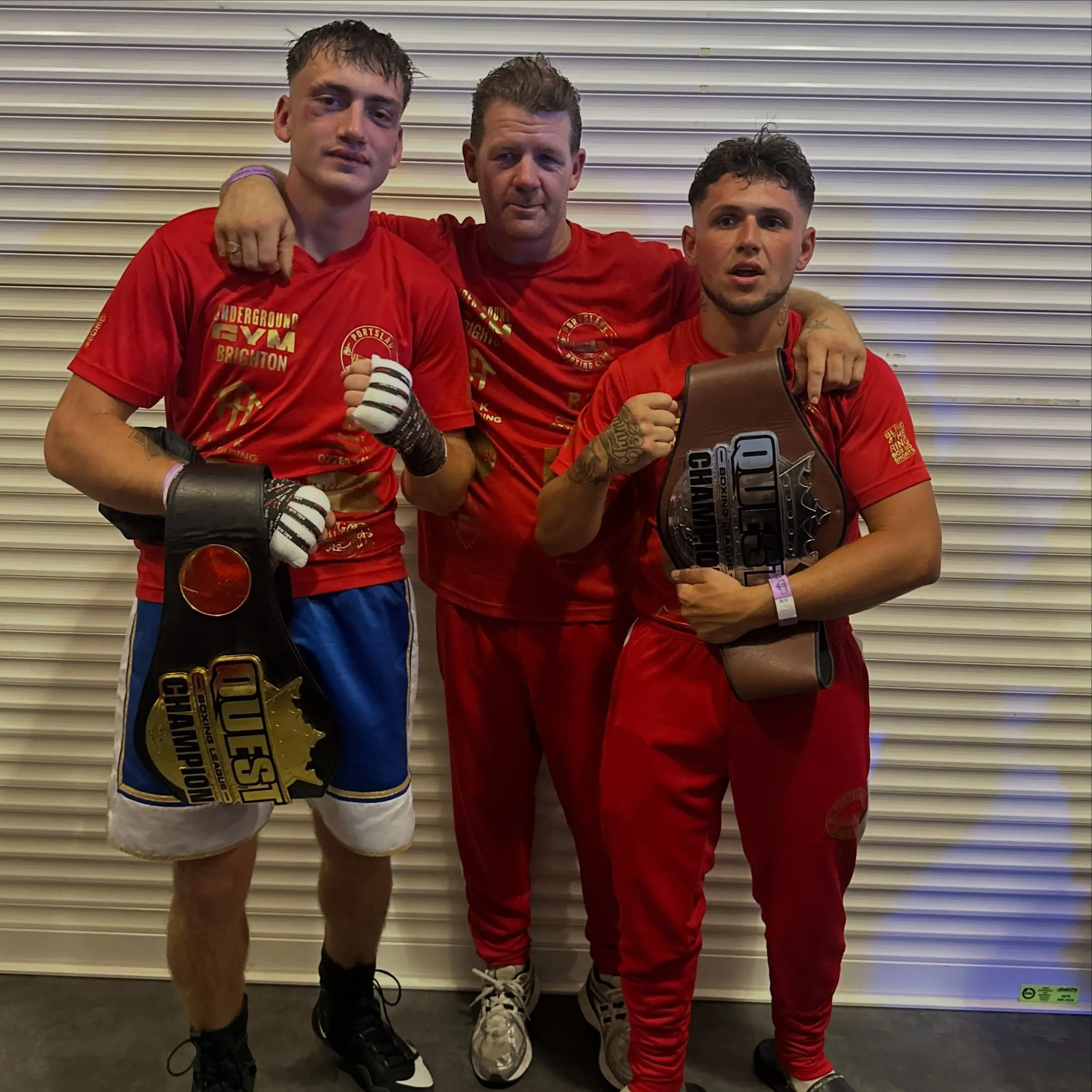 Two boxers in red holding Quest title belts with their coach