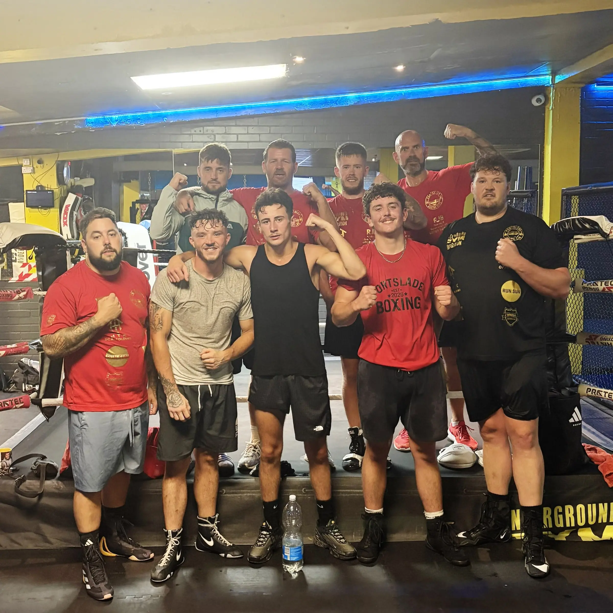 A group of boxers in red and black training kit posing together in a gym with a ring behind them